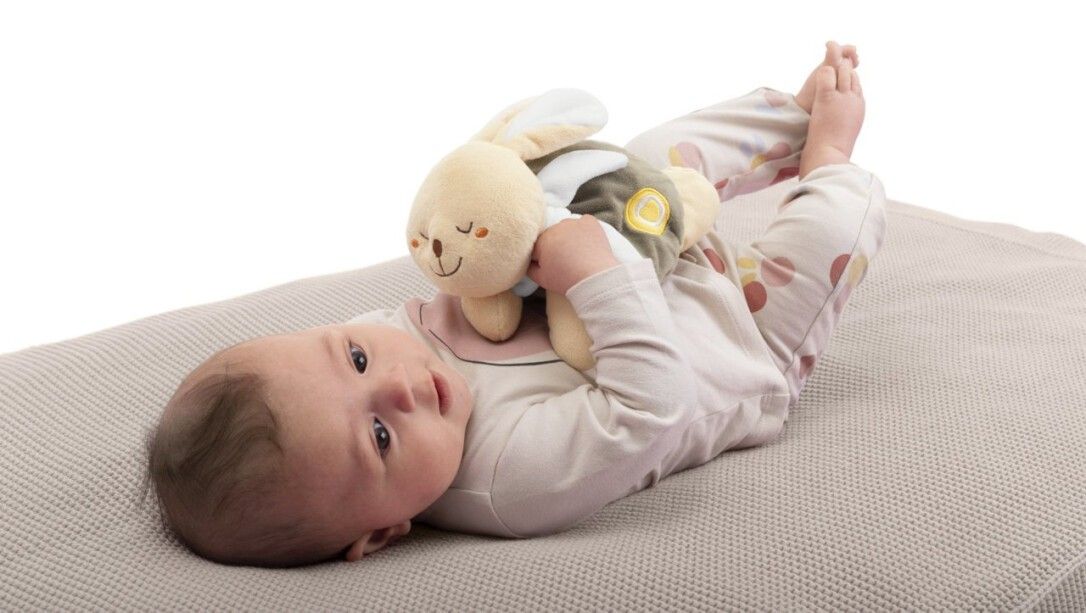 Bambino sdraiato su una coperta, tiene in mano un coniglietto di peluche beige. Il bambino guarda la fotocamera.