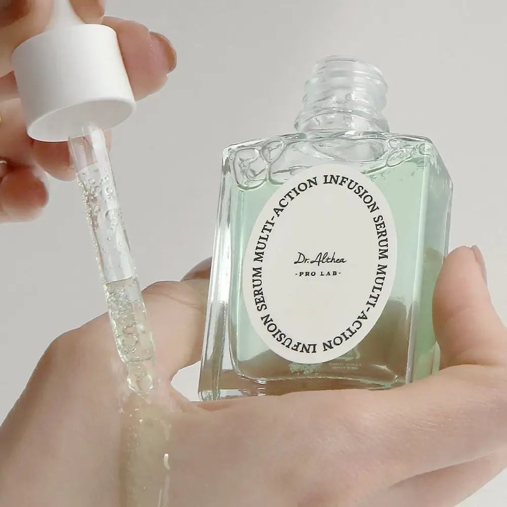 Liquido chiaro versato da una bottiglia di vetro su una mano con una pipetta. Scritta: Multi-Action Infusion Serum.