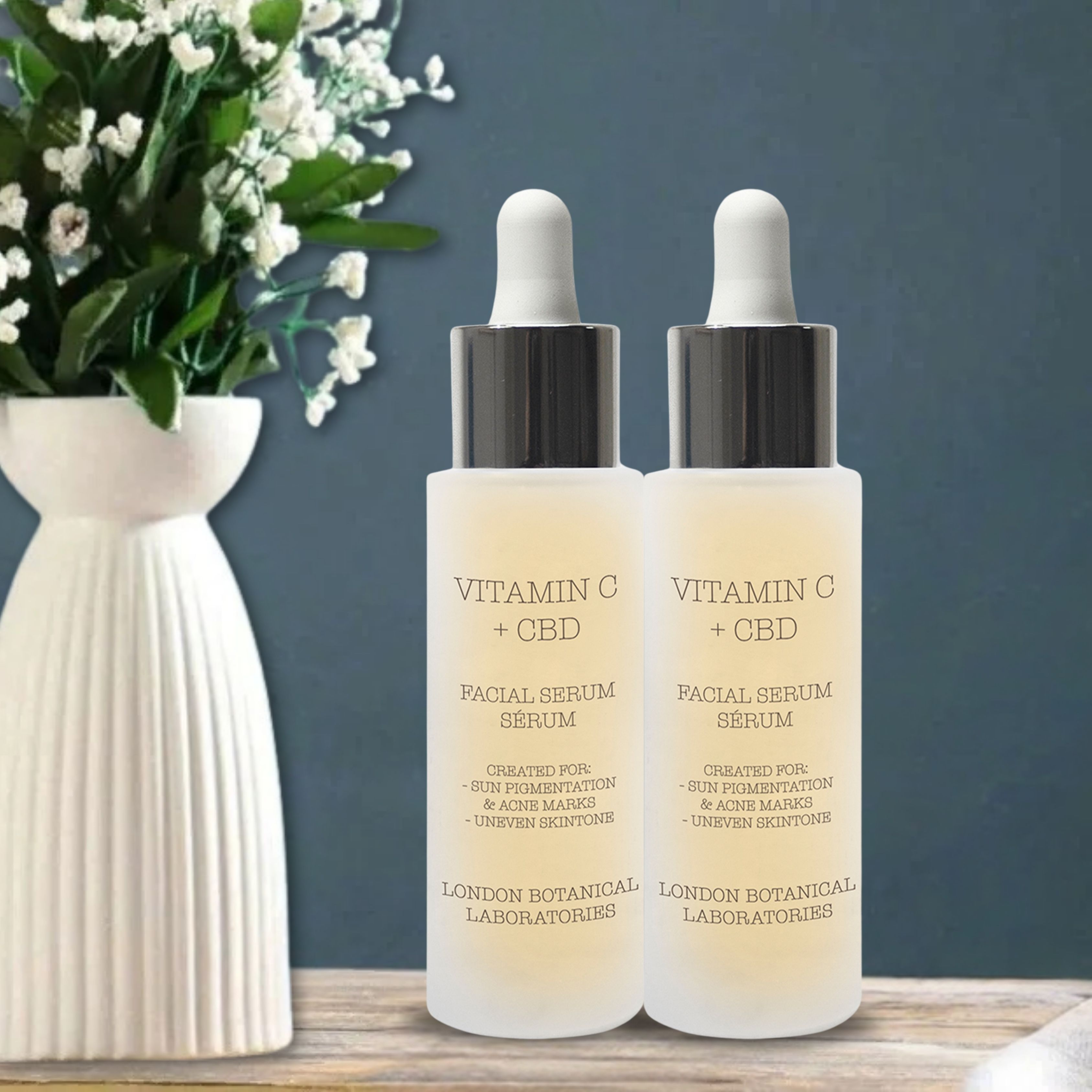 Due flaconi di siero con contagocce. Scritta: Vitamin C + CBD. Marchio: London Botanical Laboratories. Sfondo: vaso con fiori.