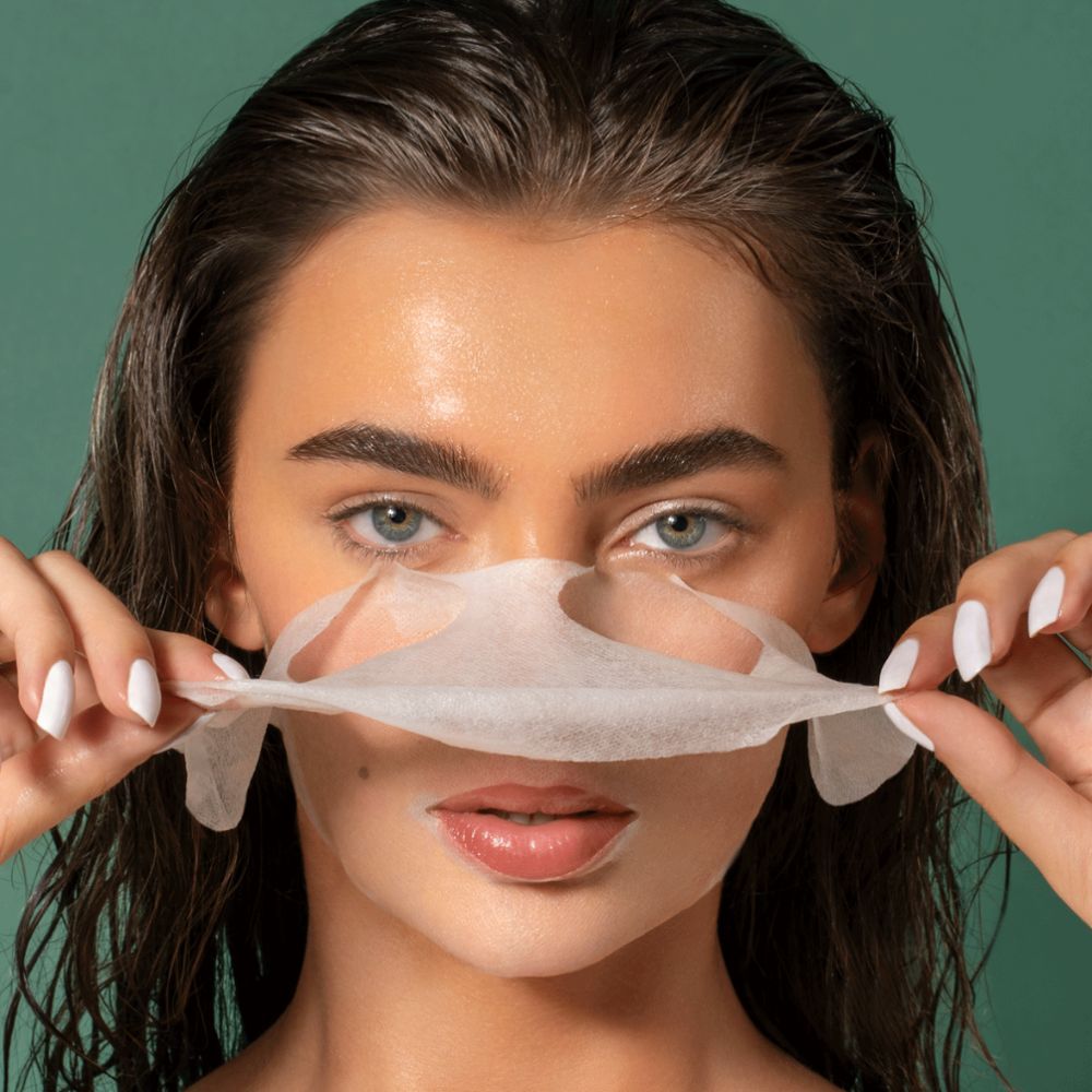 Donna tiene in mano una maschera per il viso bianca. La pelle del viso è visibile. Sfondo verde.