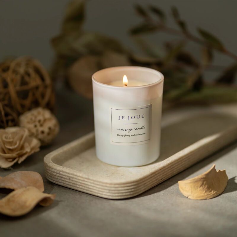 Candela bianca accesa in un contenitore di vetro. Su un vassoio. Etichetta: Je Joue, massage candle. Sfondo: decorazioni.