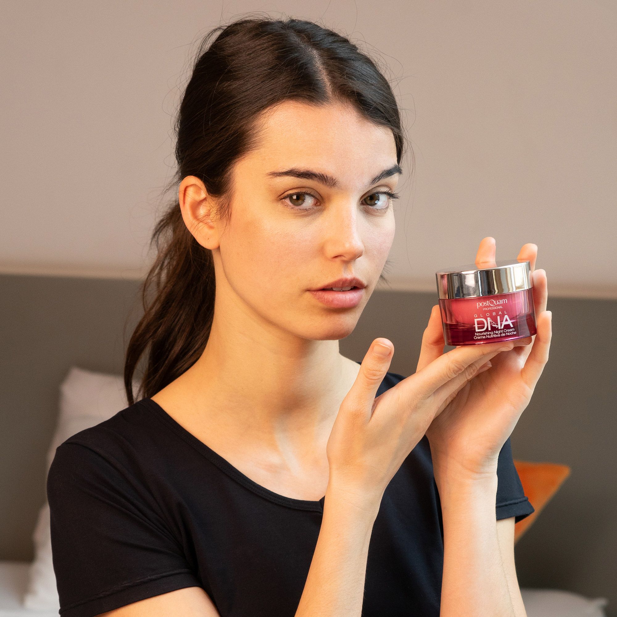 Donna che tiene un vaso. Scritta: Postquam Global DNA Nourishing Night Cream. Vaso rosa con coperchio argentato, davanti a una persona.