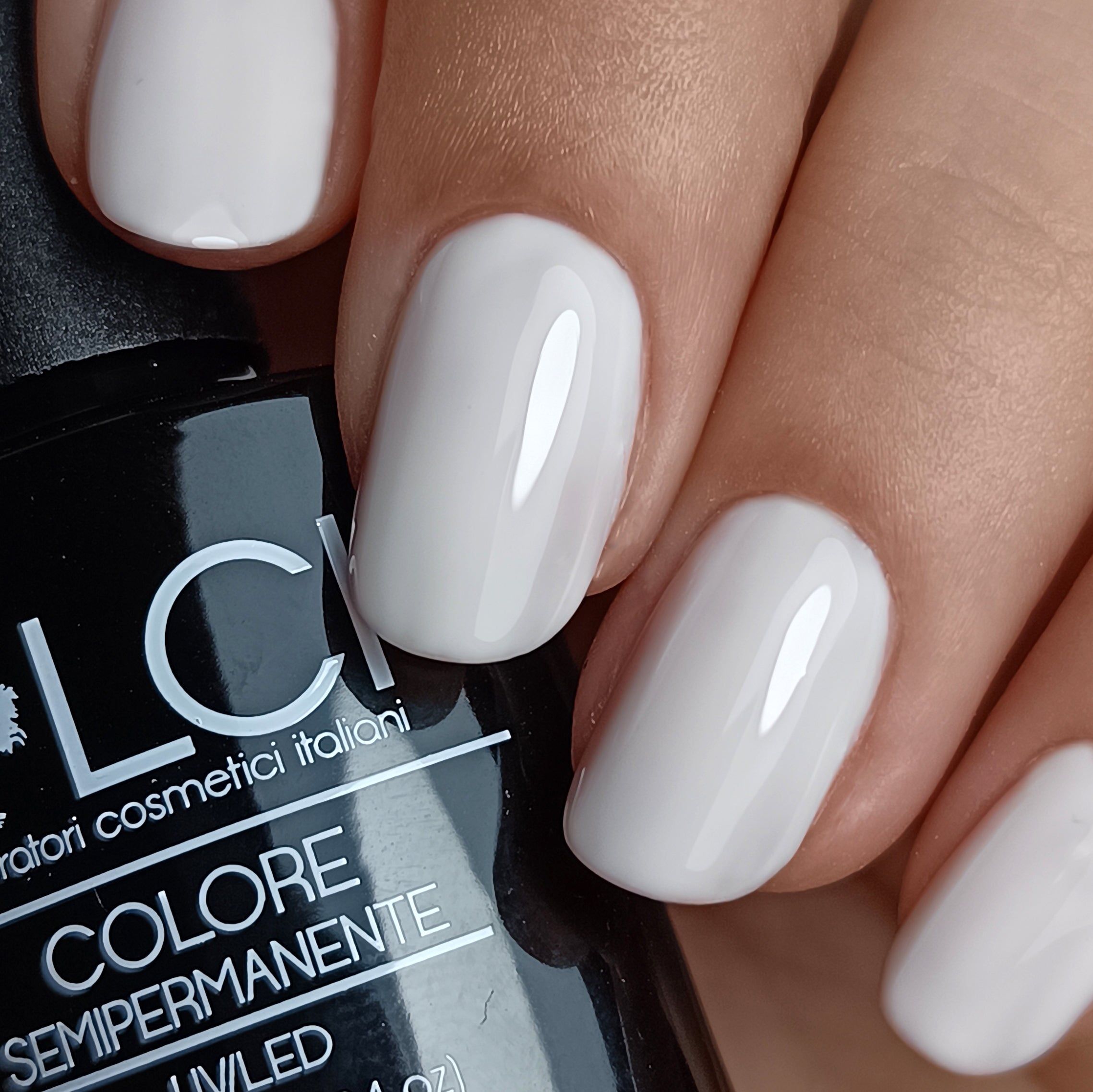 Primo piano di unghie smaltate di bianco. Flacone con scritta LCI, Colore Semipermanente sullo sfondo.