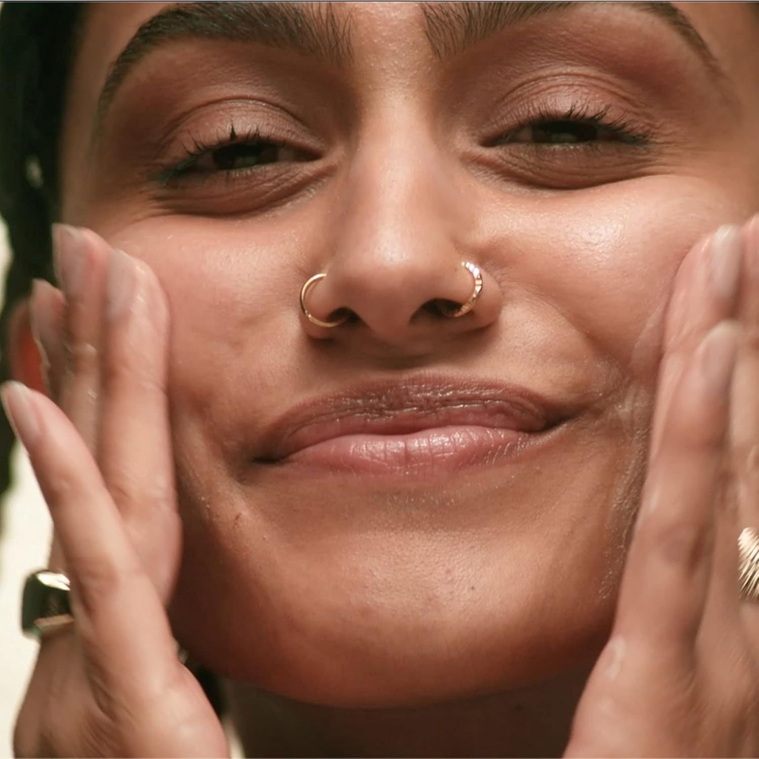 Primo piano di una persona che si mette le mani sul viso. Indossa un piercing al naso e un anello d'oro.