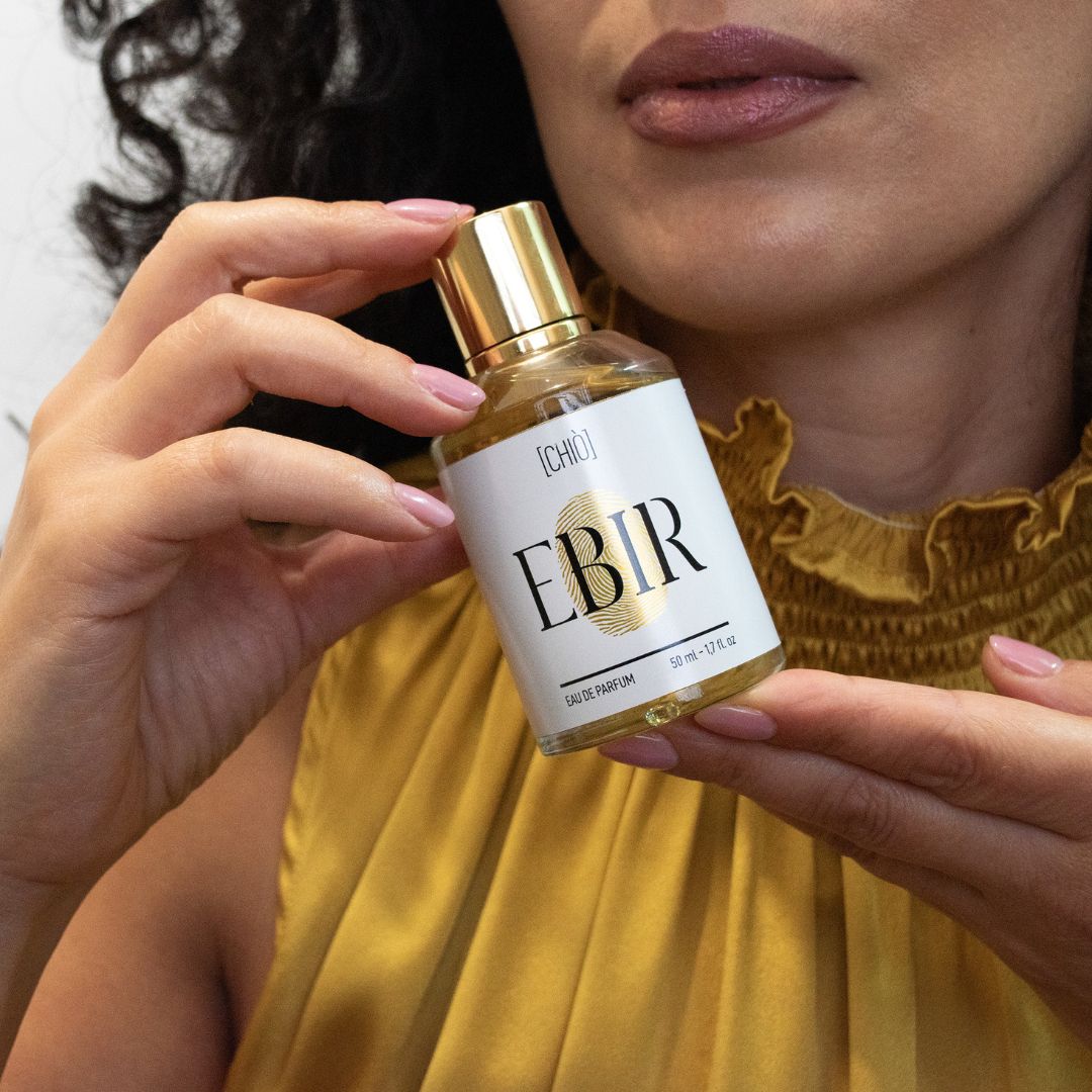 Donna tiene in mano flacone di profumo. Tappo dorato, etichetta bianca con testo nero: EBIR. Liquido dorato visibile.