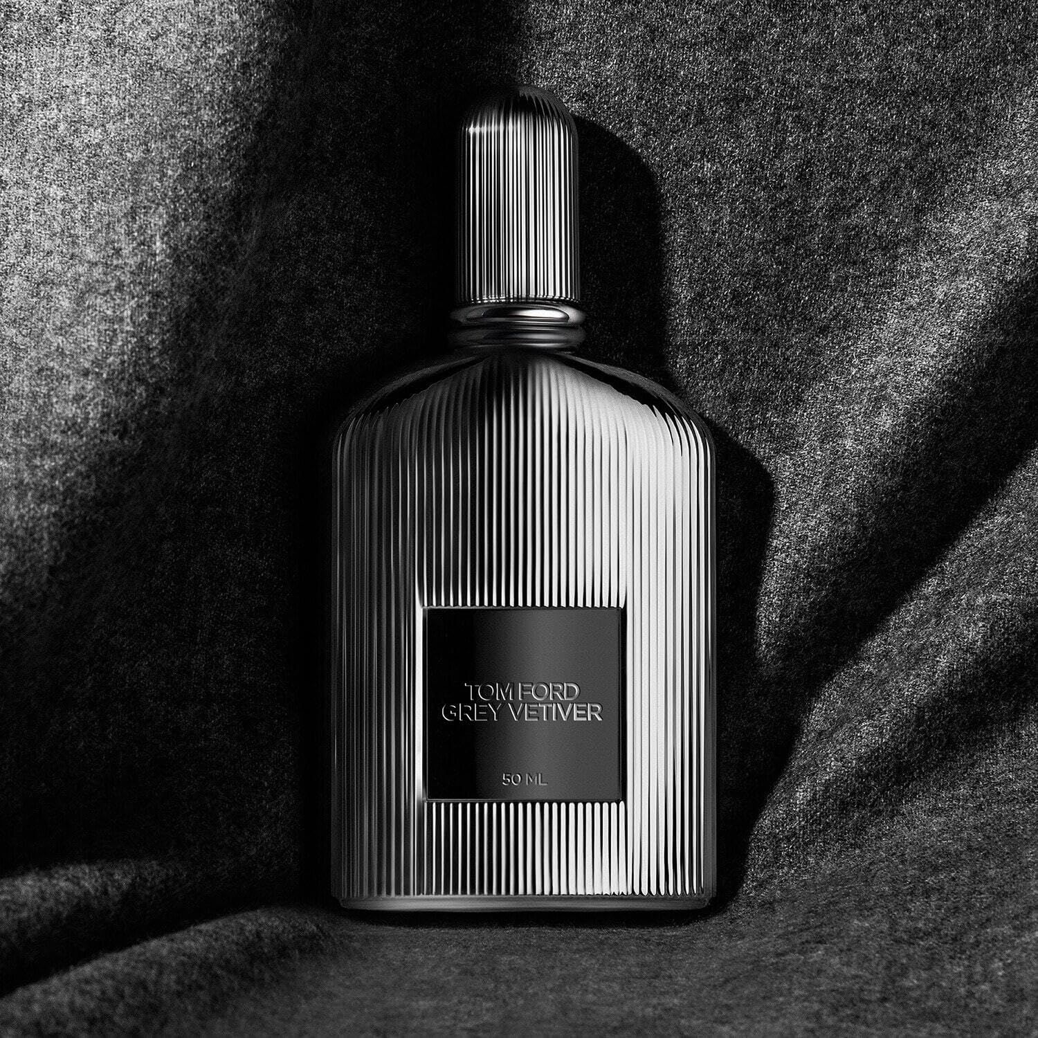 Flacone grigio con superficie a coste e tappo argentato. Etichetta rettangolare con testo: TOM FORD GREY VETIVER. Su tessuto grigio.