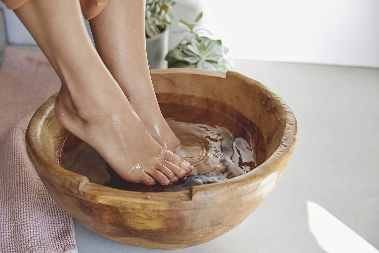 Piedi in una bacinella di legno con acqua. Pelle chiara, piedi leggermente arrossati. Sfondo: piante, asciugamano rosa, pavimento chiaro.