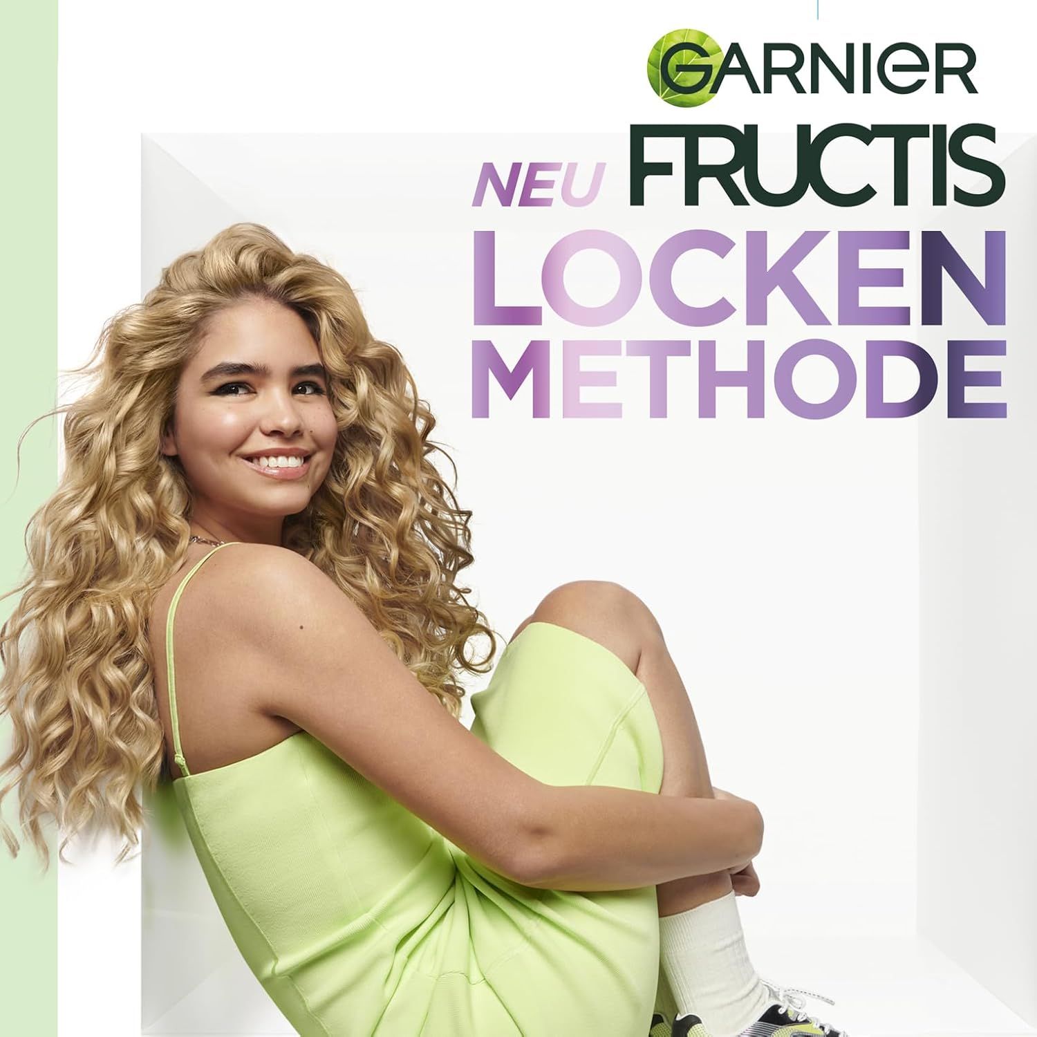 Giovane donna con capelli ricci seduta. Testo: Garnier Fructis Locken Methode. La donna indossa un vestito verde chiaro e sorride.
