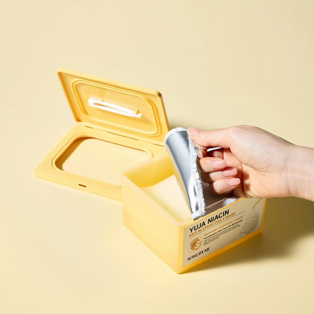 Confezione di maschere gialla aperta. Mano che estrae un involucro di maschera argentato. Scritta: Yuja Niacin Anti Blemish Daily Mask.