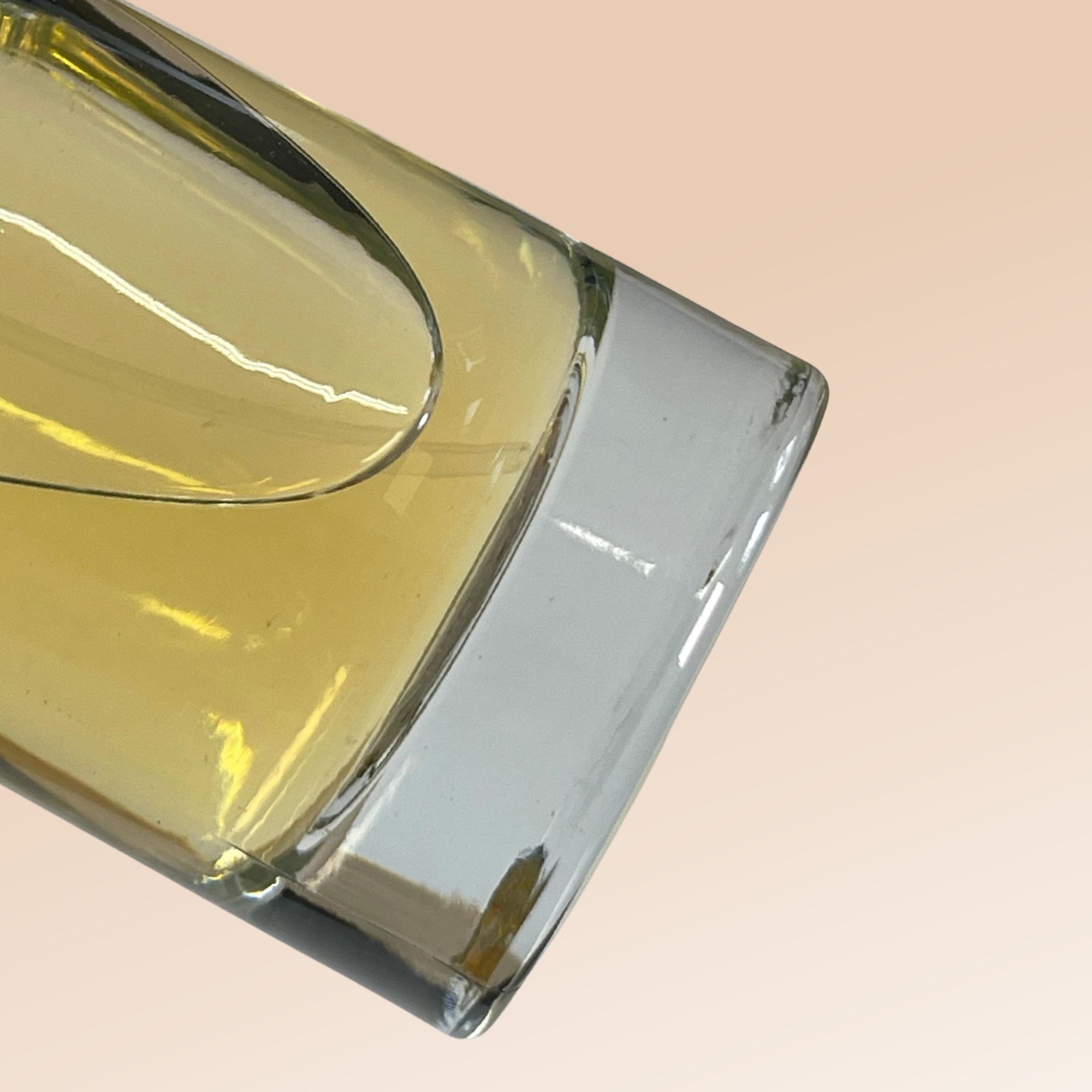 Flacone di profumo, forma quadrata, vetro trasparente. Liquido dorato all'interno. Base rettangolare grigia.