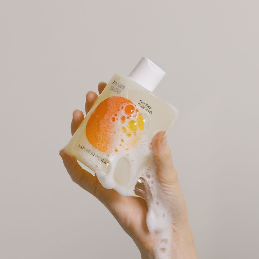 Una mano tiene in mano un flacone di bagnoschiuma. La schiuma fuoriesce dal flacone. Sul flacone sono visibili testo e un disegno arancione.