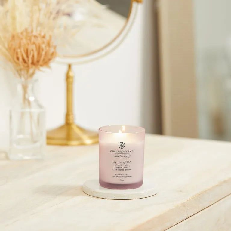 Candela rosa Chesapeake Bay mind & body, joy + laughter, cranberry dahlia, su sottobicchiere. Specchio e vaso sullo sfondo.