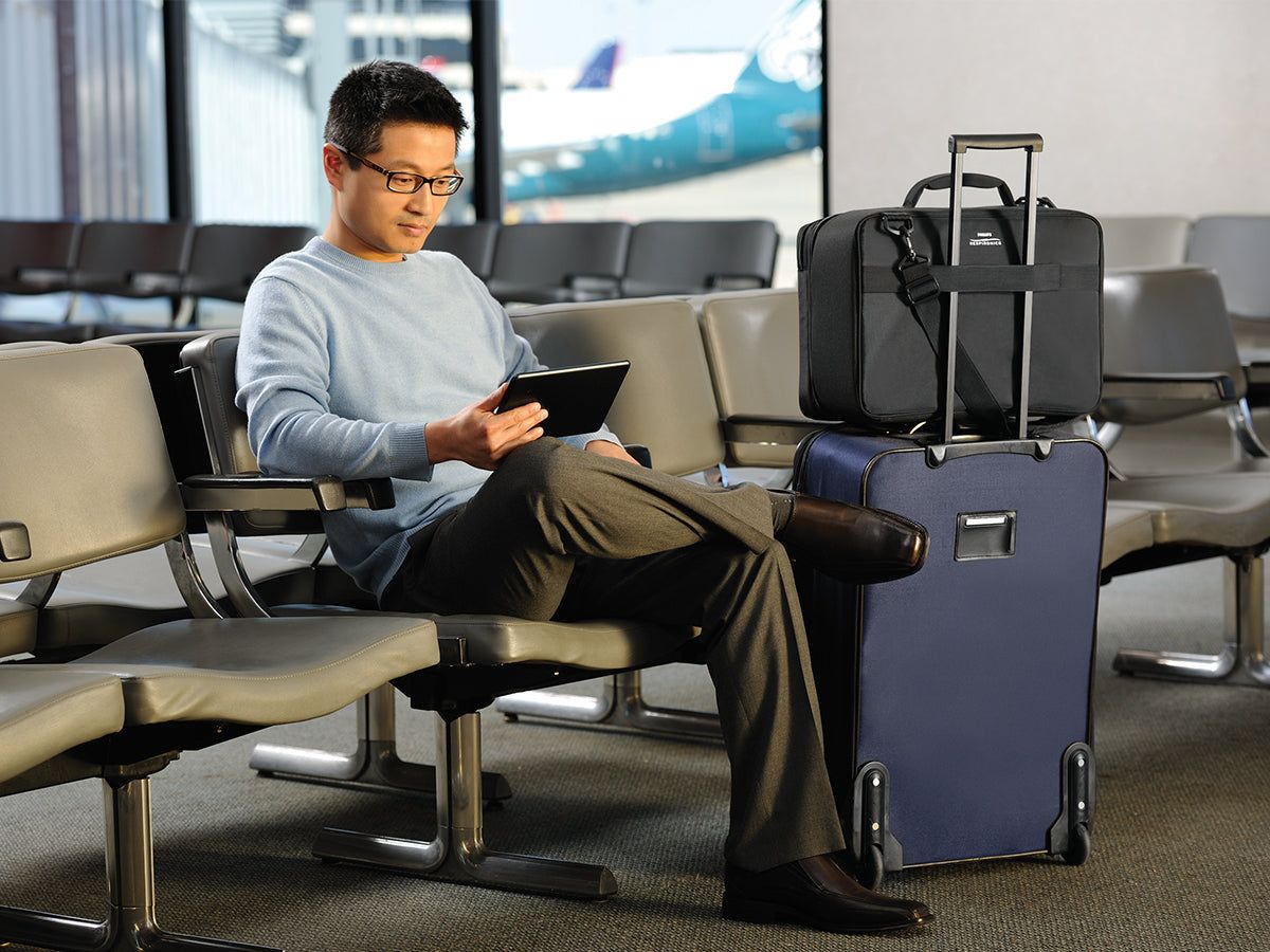 Uomo seduto in una sala d'attesa dell'aeroporto con una borsa da viaggio nera su una valigia con ruote accanto a lui.