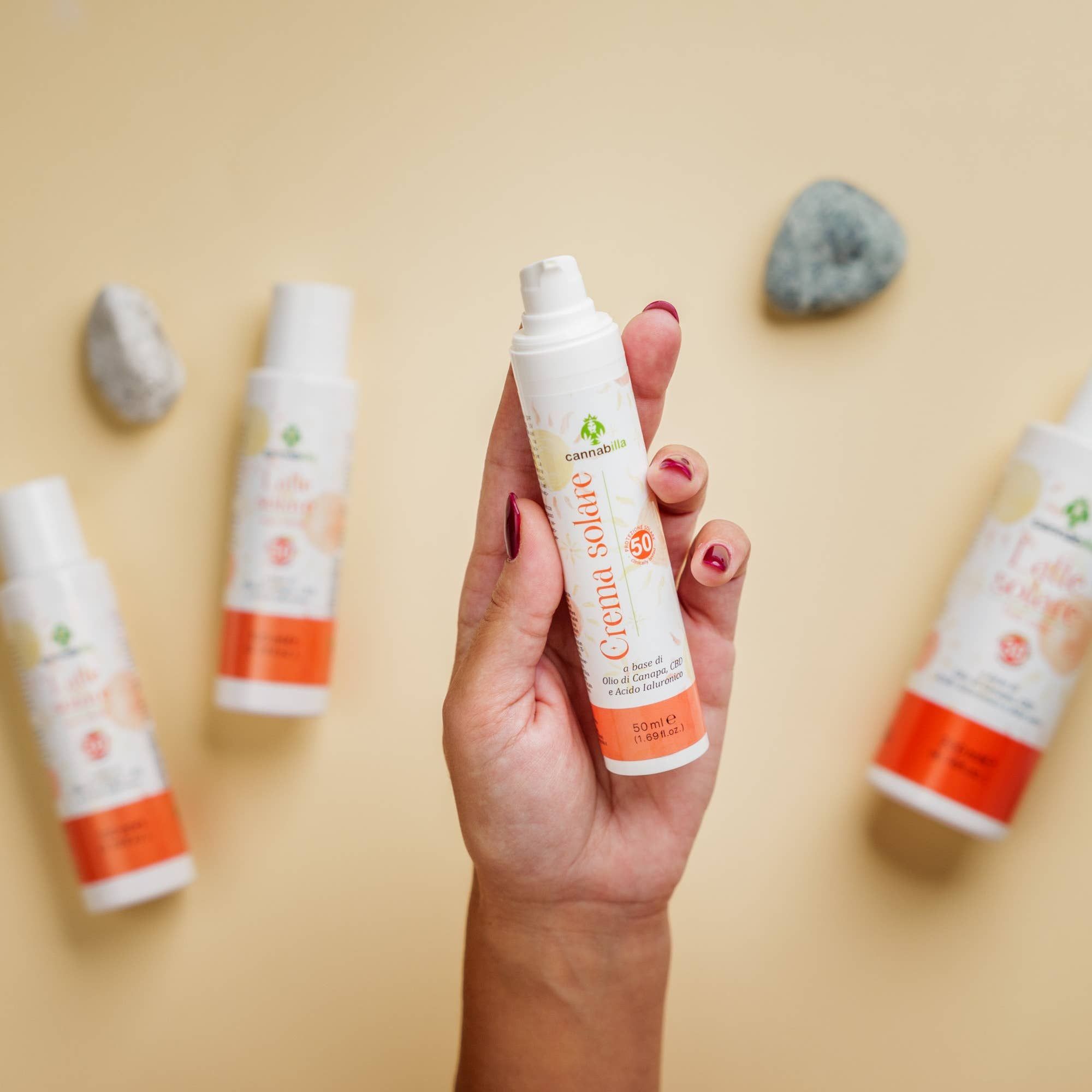 Mano che tiene un flacone bianco con dosatore. Scritta: Crema solare, SPF50. Marchio: Cannabilla. Diversi flaconi sullo sfondo.
