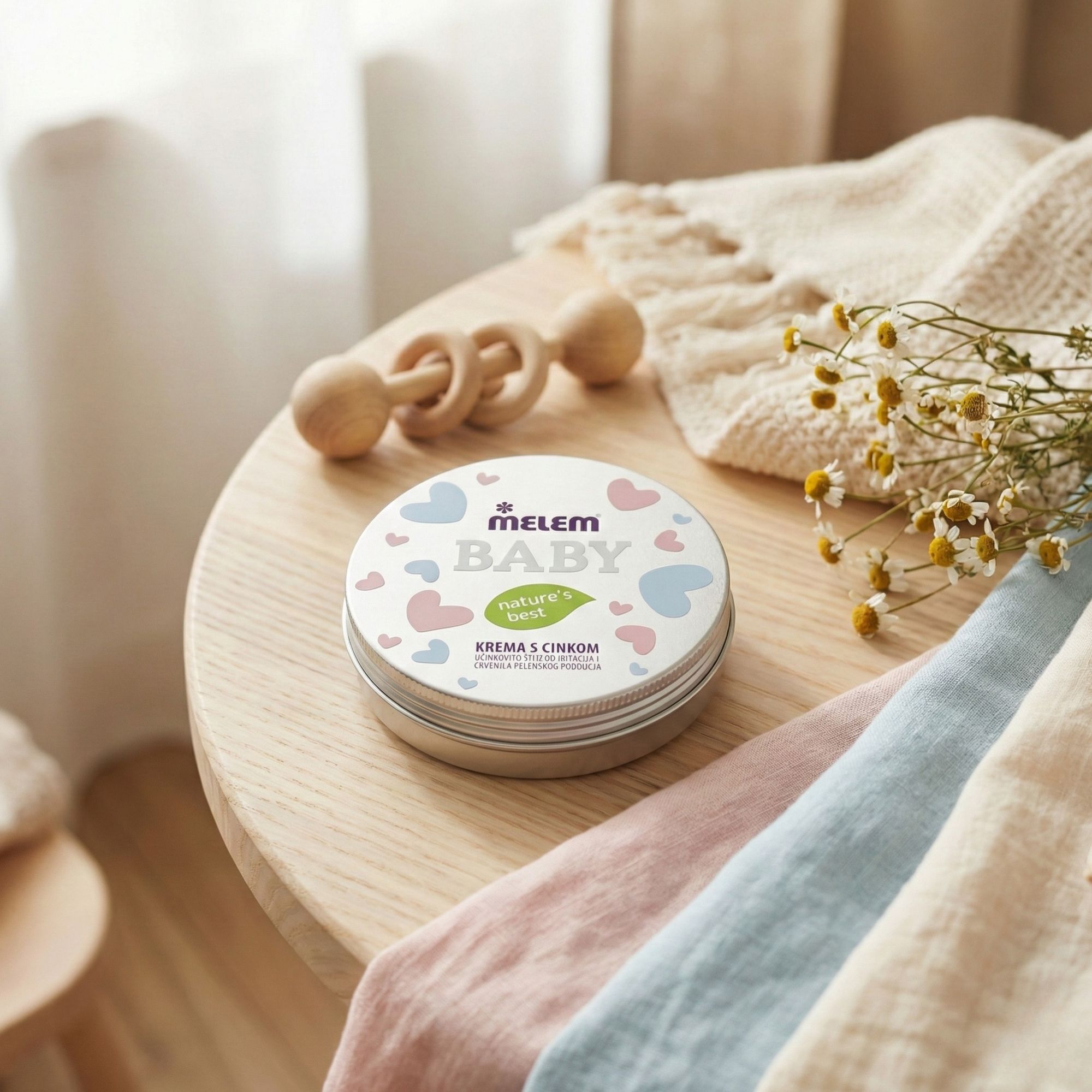 Barattolo rotondo Melem Baby su un tavolo con un sonaglio di legno e pannolini. Fiori di camomilla.