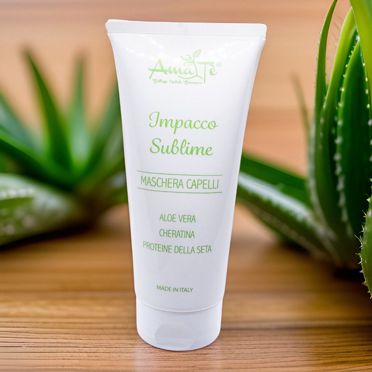 Tubo bianco con testo verde: Amatè, Impacco Sublime, Maschera Capelli. Aloe Vera, Cheratina, Proteine della seta. Su legno, con piante di Aloe Vera sullo sfondo.