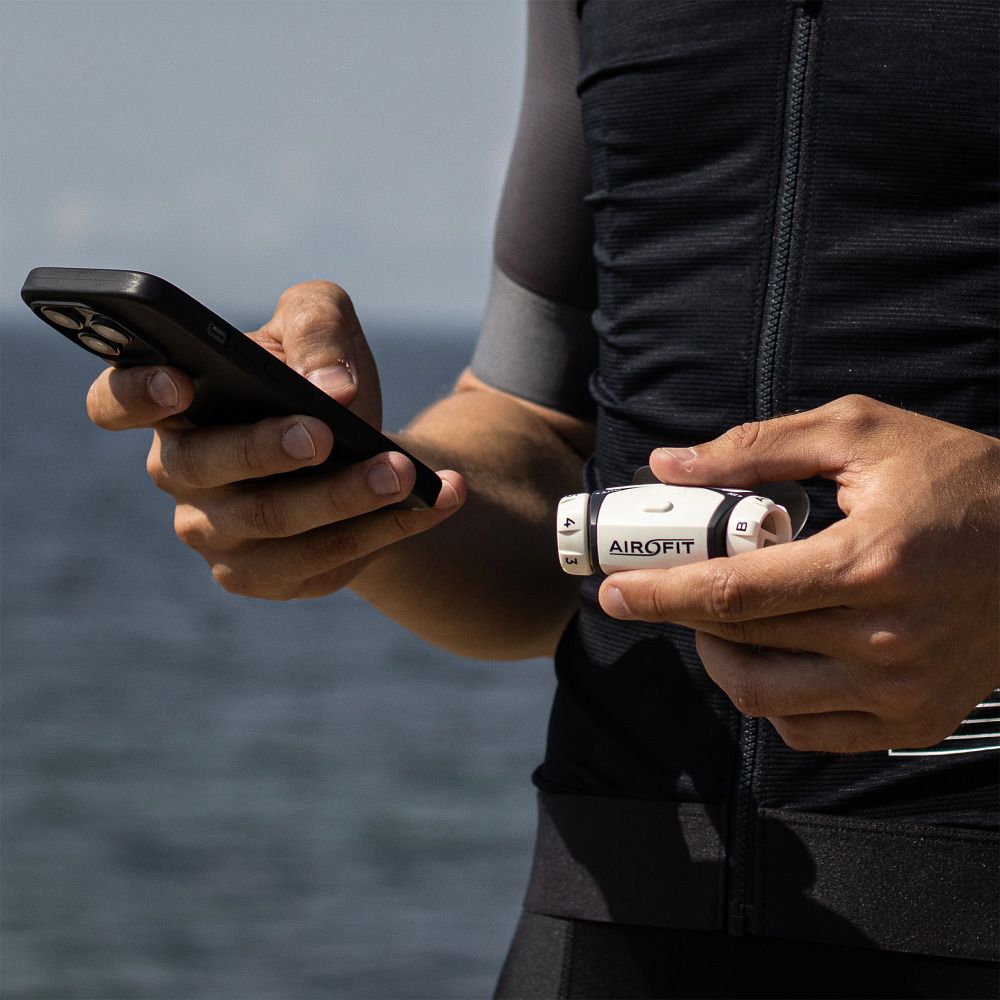 Persona tiene smartphone e dispositivo di allenamento respiratorio bianco con logo Airofit. Dettagli neri e scala.