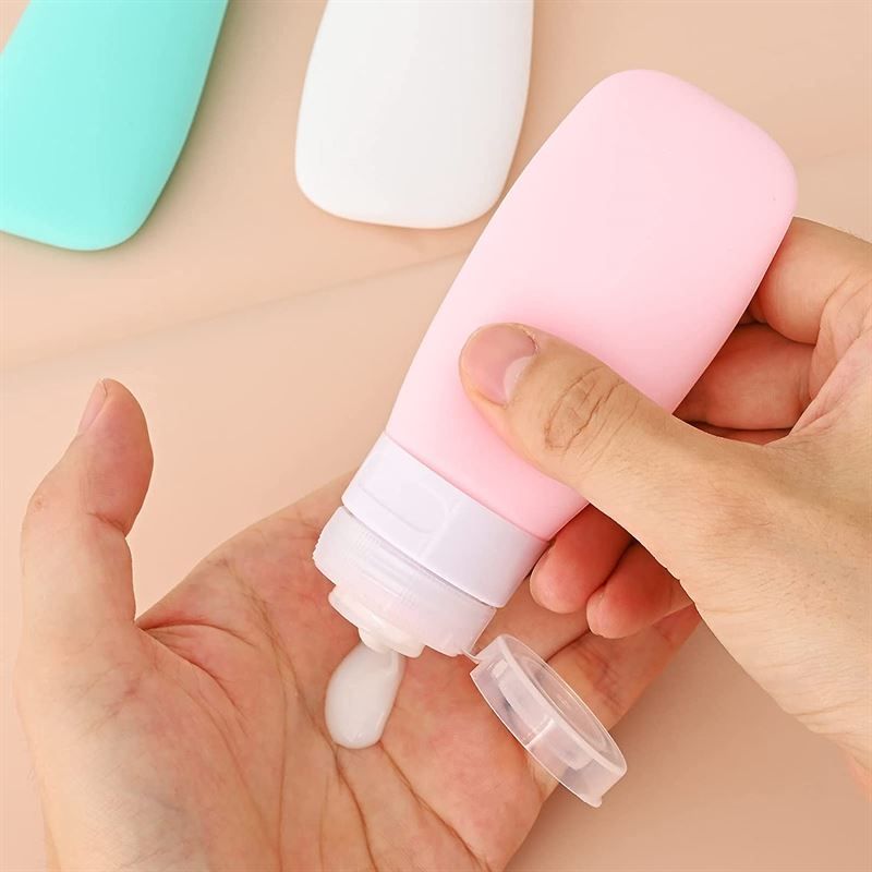Flacone da viaggio rosa con coperchio bianco. Una mano tiene il flacone, da cui esce un liquido. Coperchio accanto.