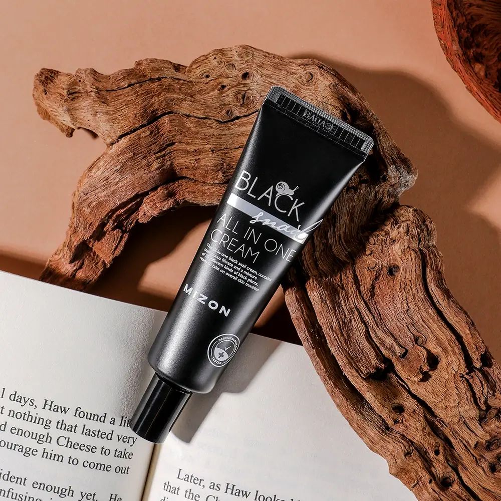 Tubo nero con scritta BLACK Snail ALL IN ONE CREAM e MIZON. Su legno e libro.