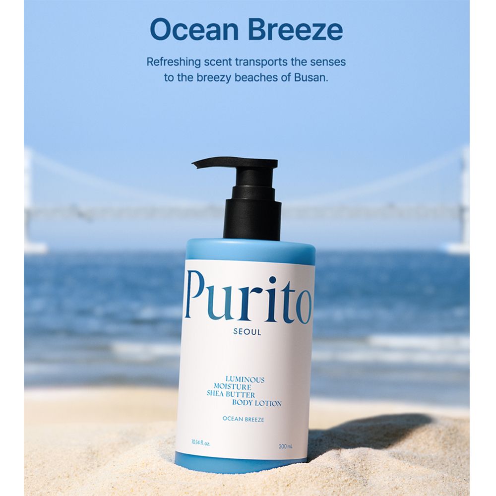 Flacone blu chiaro con erogatore nero sulla sabbia. Scritta: Purito Seoul, Luminous Moisture Shea Butter Body Lotion, Ocean Breeze.