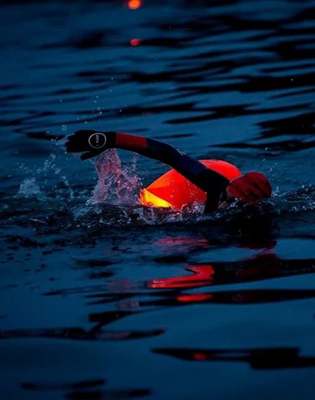 Persona che nuota in acqua con boa arancione. Ambiente scuro, l'acqua riflette la luce.