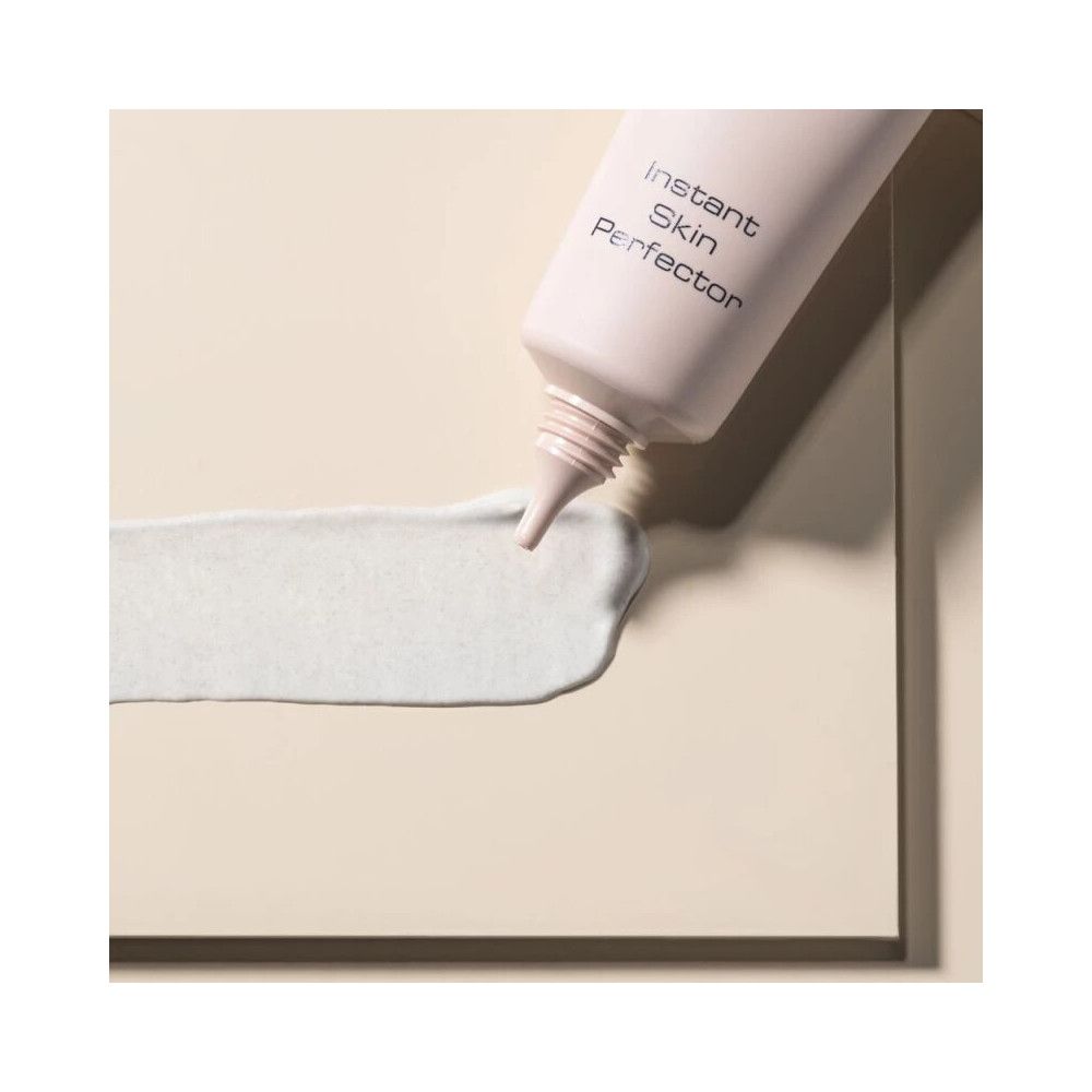 Tubo di prodotto e macchia. Scritta Instant Skin Perfector. Tubo rosa, punta. Macchia verde chiaro. Sfondo beige.