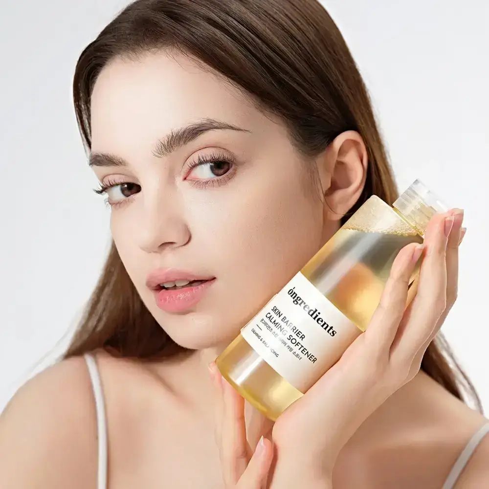 Una donna tiene in mano una bottiglia trasparente con liquido giallastro. L'etichetta dice "ongredients SKIN BARRIER CALMING SOFTENER".