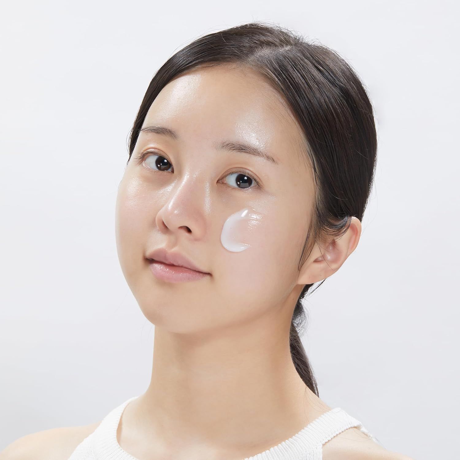 Primo piano di una donna che applica la crema sul viso. Pelle chiara, capelli scuri. Canotta bianca.