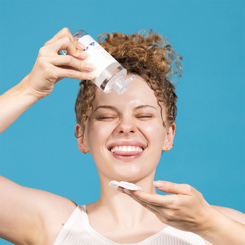 Donna con occhi chiusi che tampona un batuffolo di cotone con liquido. Sfondo blu. Bottiglia visibile.