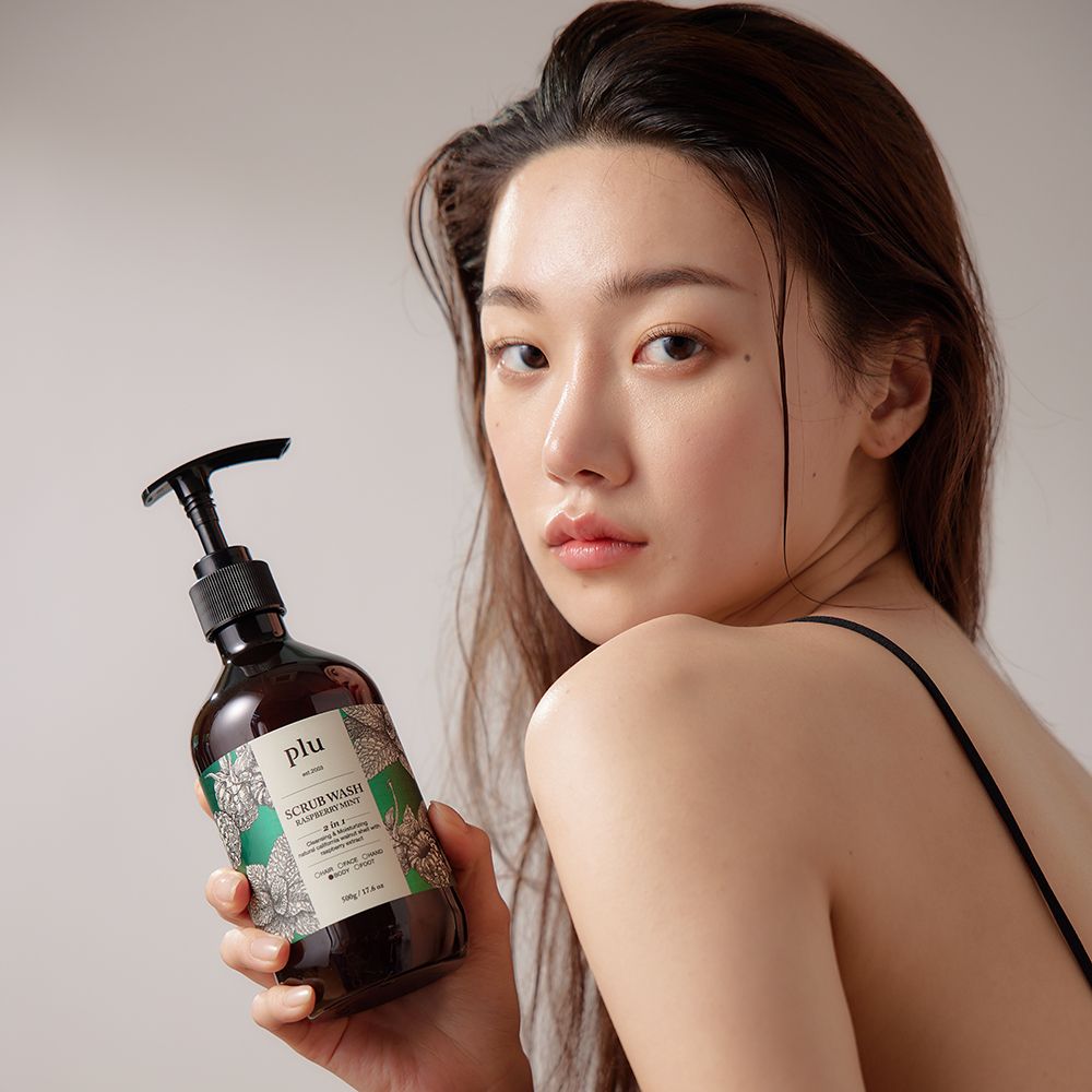 Una donna tiene in mano una bottiglia marrone con erogatore nero. Scritta: PLU Scrub Wash Raspberry Mint.