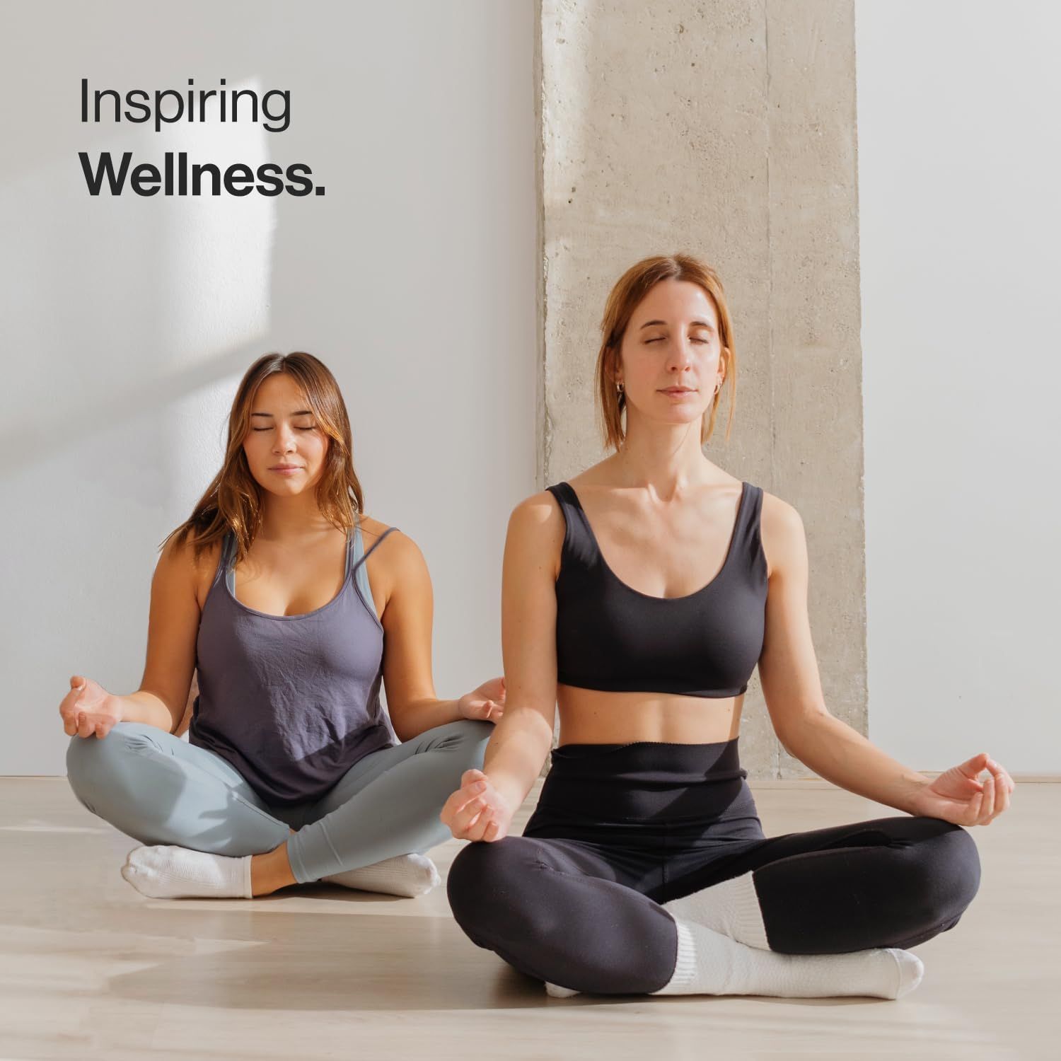 Due donne sedute a gambe incrociate meditano. Indossano abiti sportivi. Sfondo: muro di cemento.