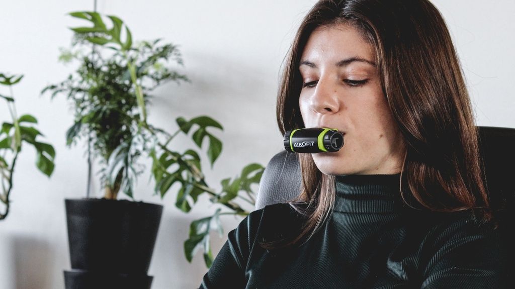 Donna che si allena con un allenatore respiratorio. Dispositivo nero-verde in bocca. La donna è seduta davanti a una pianta.