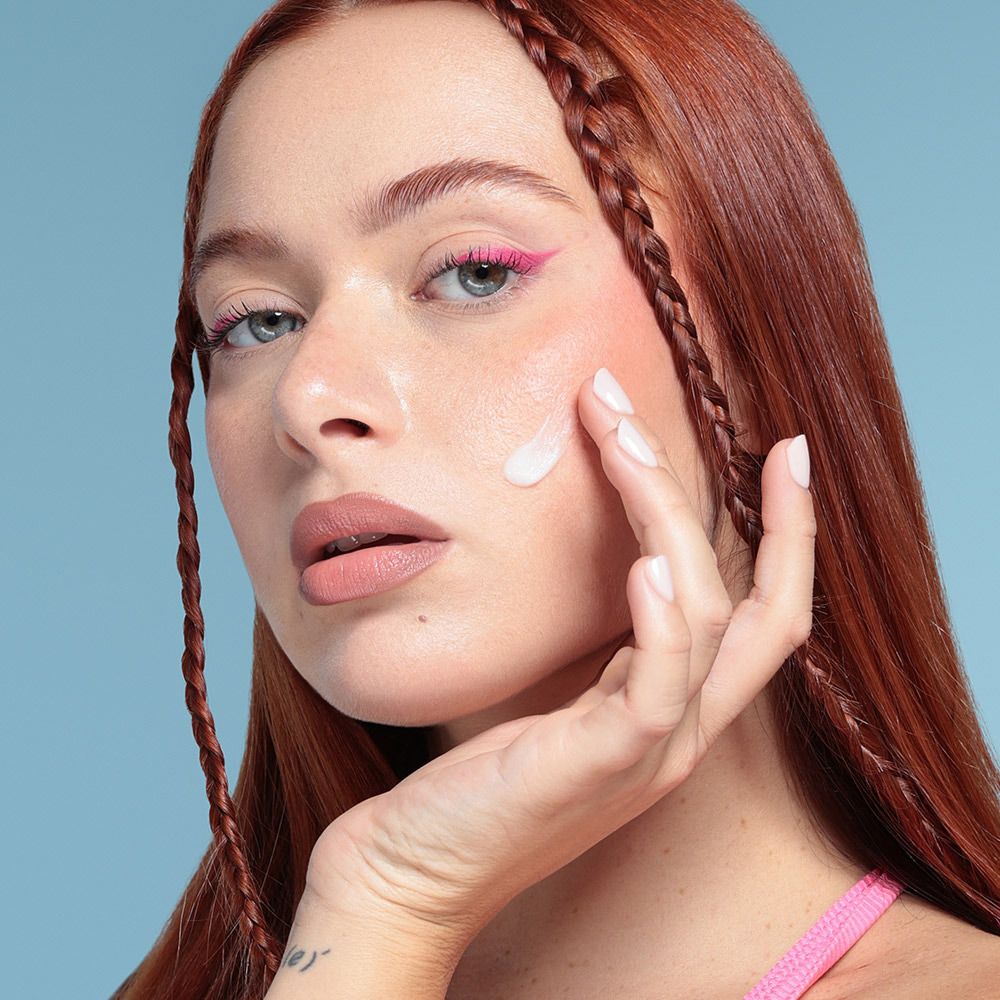 Donna applica Mia Makeup Pore Star Primer sulla guancia. Persona con trecce rosse e top rosa. Prodotto in fase di applicazione.