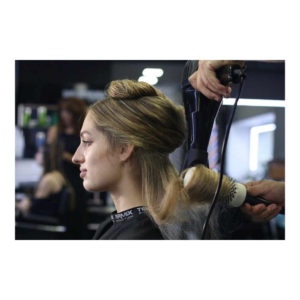 Parrucchiere asciuga i capelli di una donna con una spazzola rotonda. Altre persone sullo sfondo.