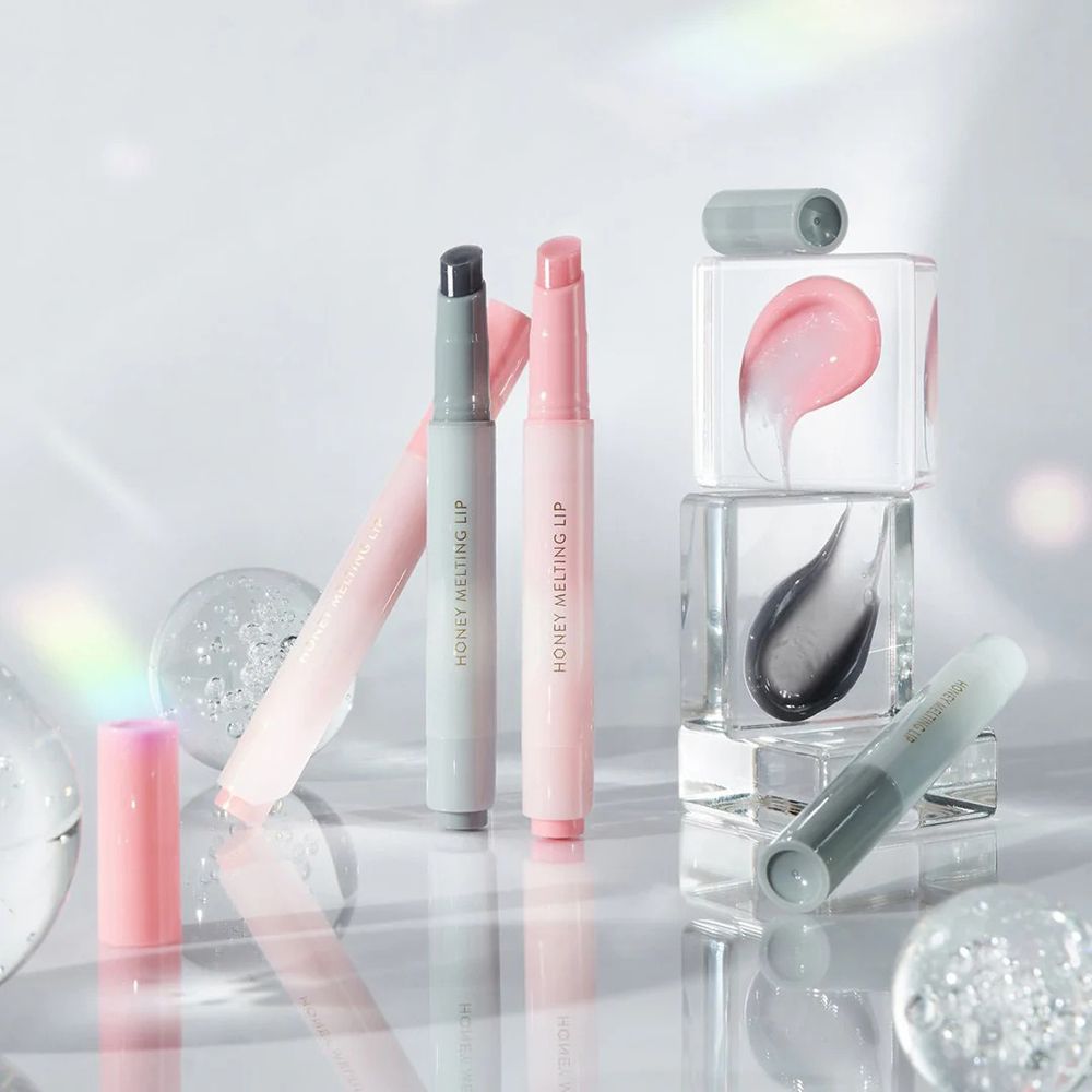 Due stick gloss, uno rosa, uno grigio-verde. Testo: HONEY MELTING LIP. Su cubi e sfere trasparenti.