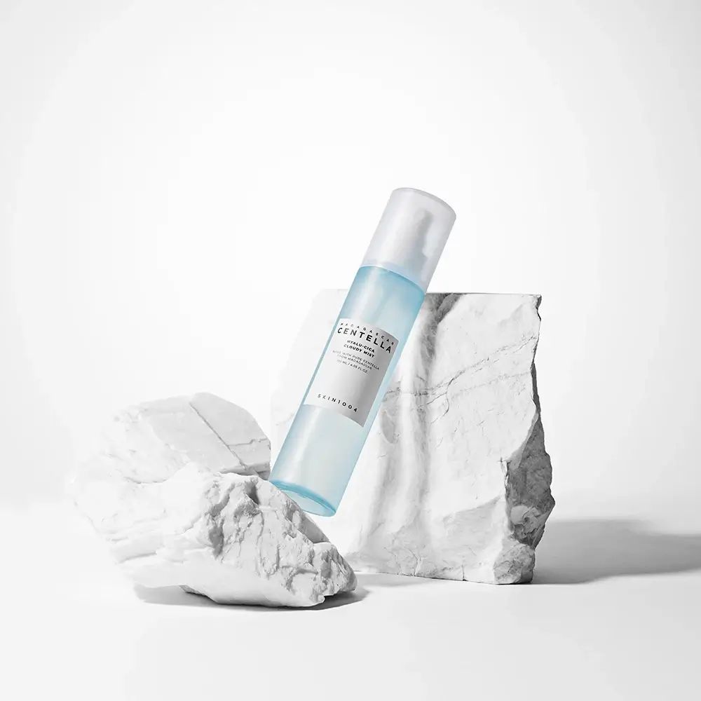 Flacone spray blu trasparente su pietre bianche. Scritta: MADAGASCAR CENTELLA, HYALU-CICA Cloudy Mist. SKIN1004.