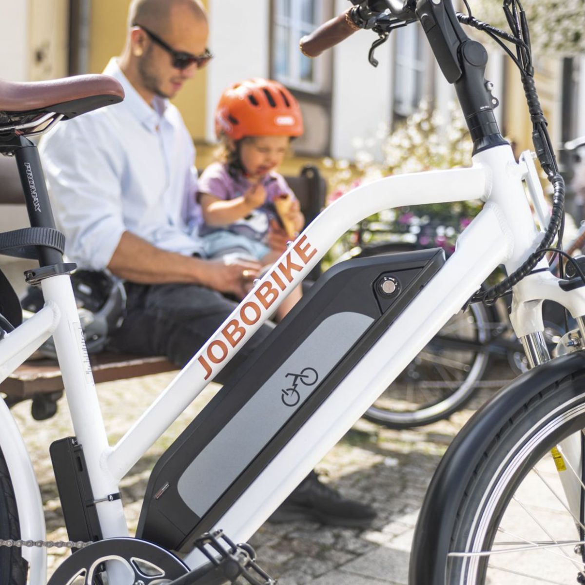 E-bike bianco con batteria, logo JOBOBIKE. Uomo e bambino seduti su una panchina sullo sfondo.