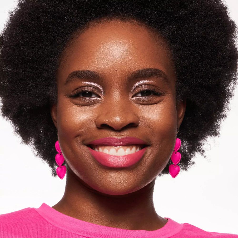 Ritratto di una donna con la pelle scura e rossetto rosa. Indossa orecchini rosa e un top rosa.