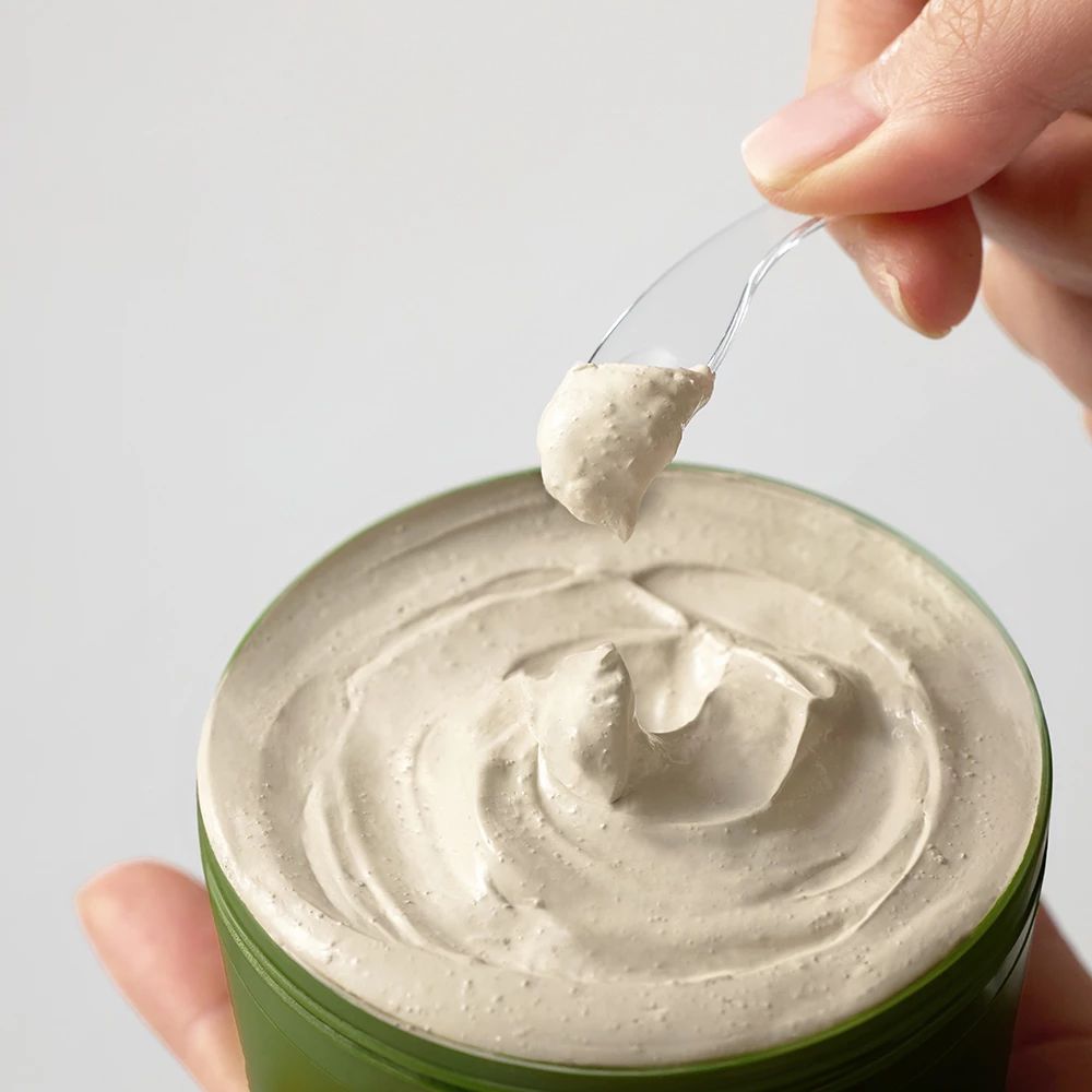 Una mano tiene una piccola spatola con una sostanza cremosa e chiara sopra un contenitore verde aperto con la stessa crema.