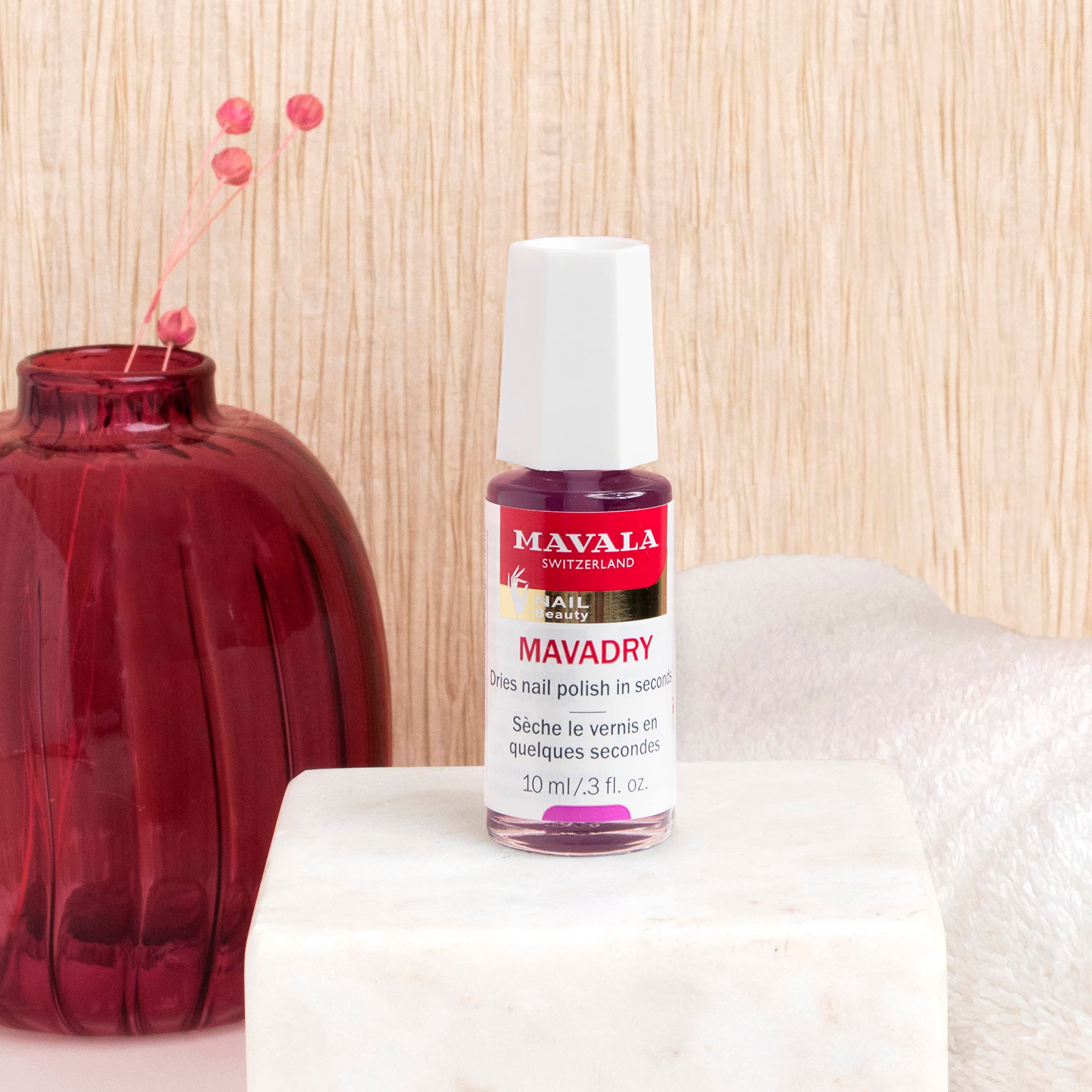 Piccola bottiglia su piedistallo bianco. Tappo rosso, etichetta con logo e nome del prodotto. Sfondo con vaso e asciugamano.