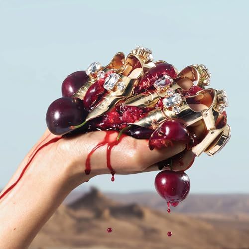 Mano con anelli dorati, tiene prugne e liquido rosso. Sfondo: deserto.