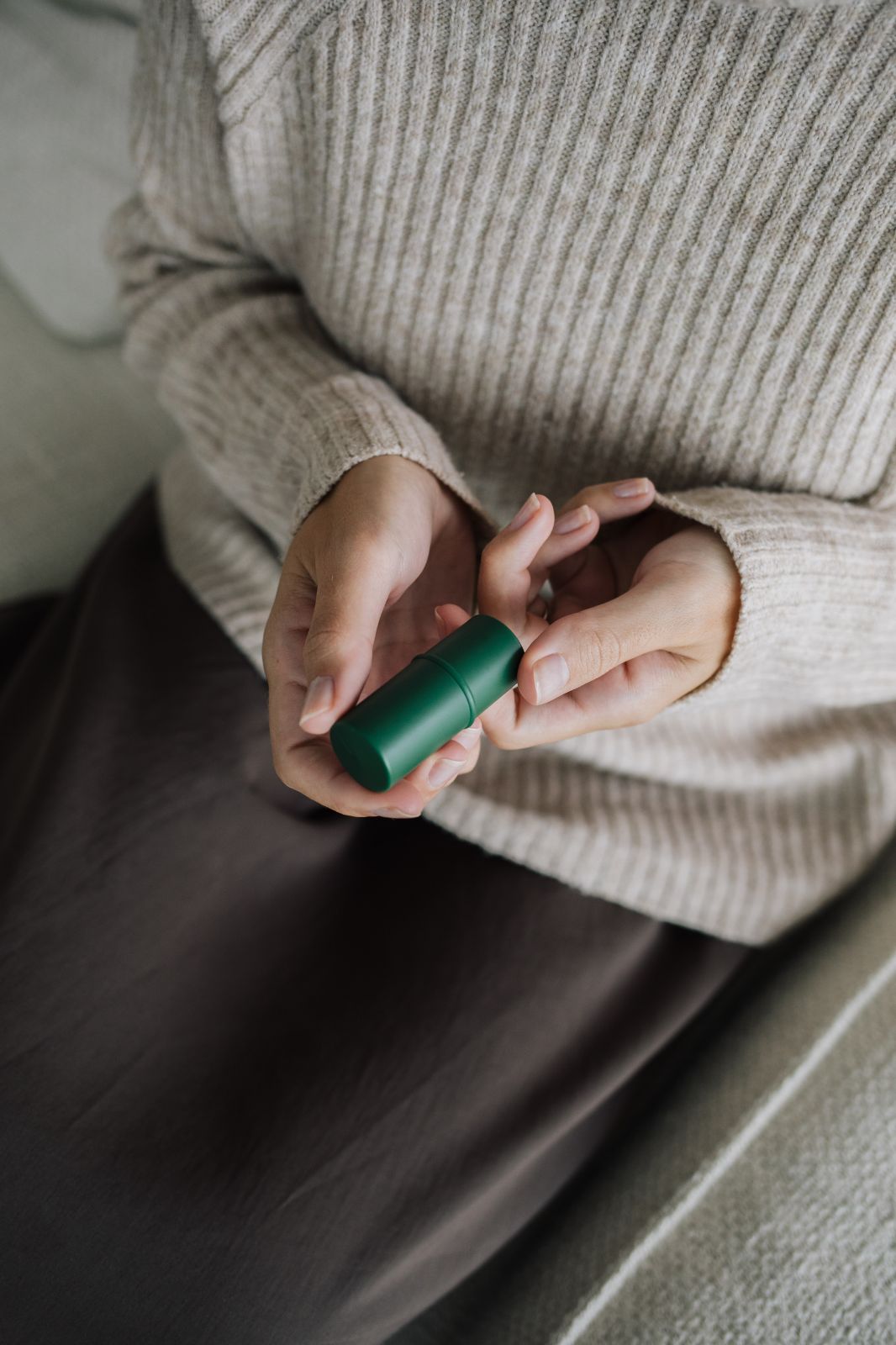 Mani che tengono uno stick di balsamo per labbra verde. La persona indossa un maglione beige e una gonna marrone.