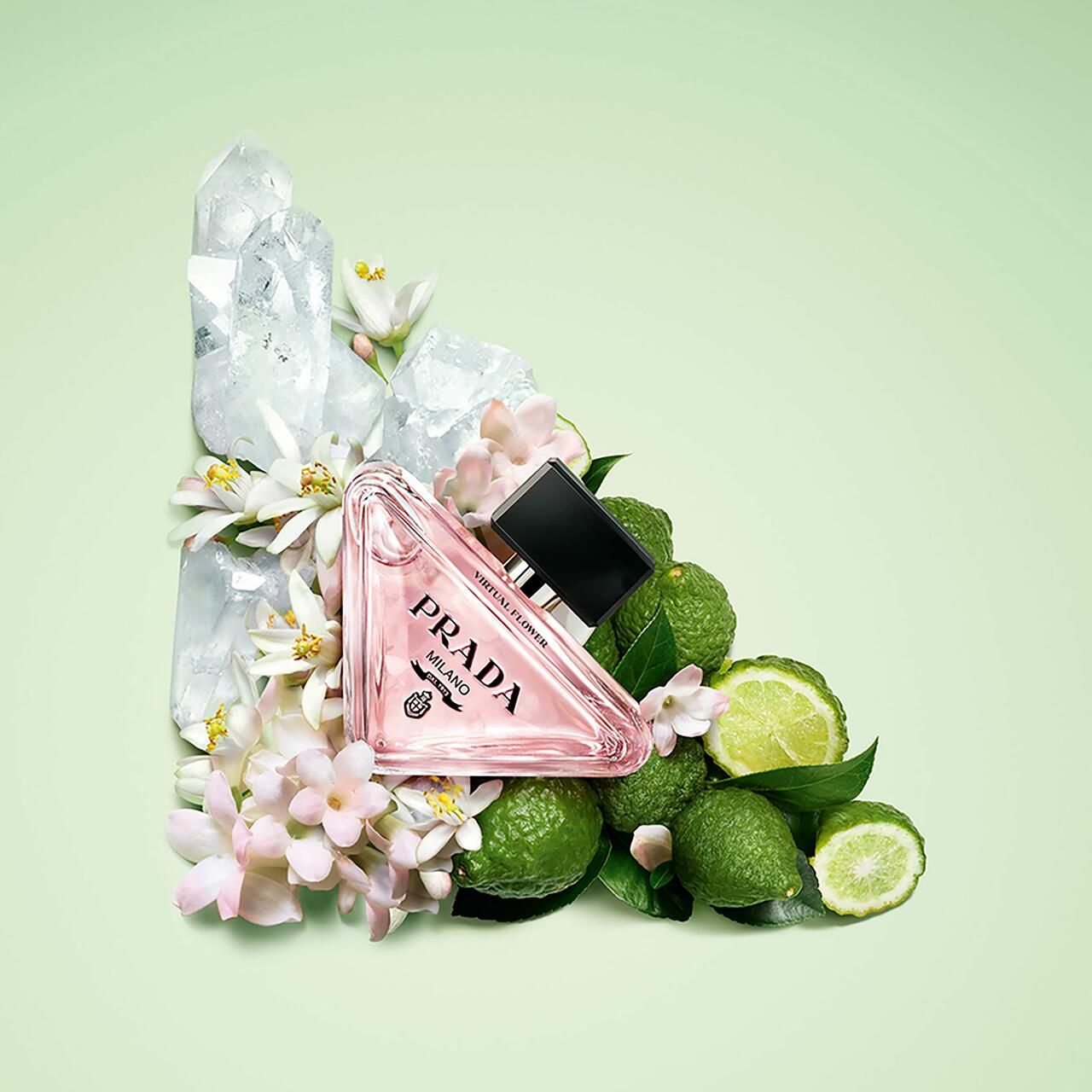 Flacone di profumo rosa con tappo nero, circondato da lime verdi, fiori e cristalli. Logo Prada.