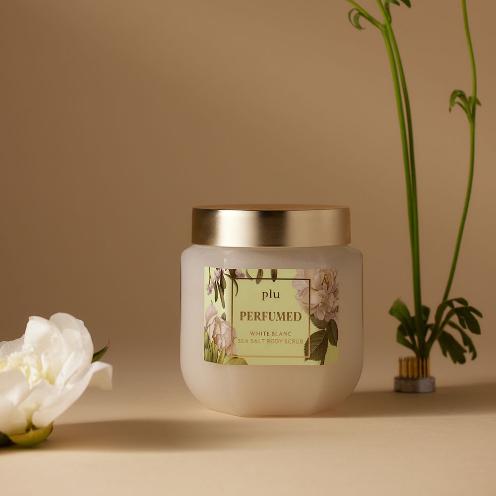Barattolo di vetro con coperchio dorato ed etichetta. L'etichetta recita "PLU PERFUMED White Blanc Sea Salt Body Scrub". Fiore bianco.