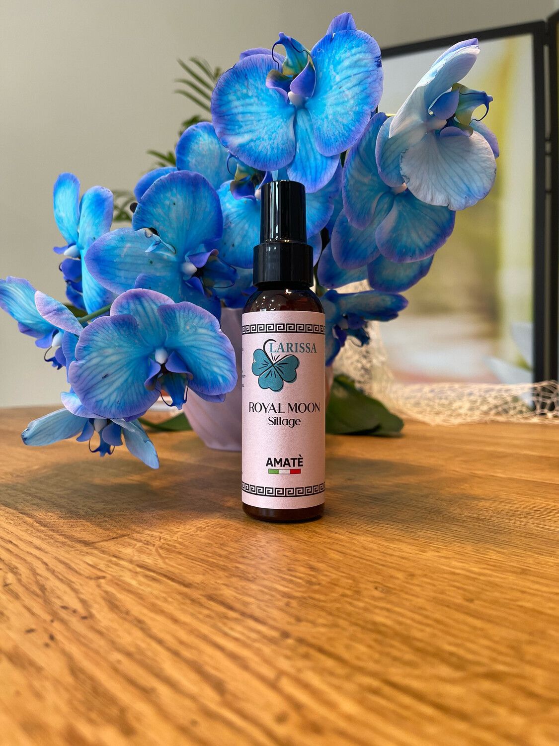 Flacone marrone con vaporizzatore nero. L'etichetta riporta "Larissa Royal Moon Sillage" e "Amatè". Davanti a orchidee blu.