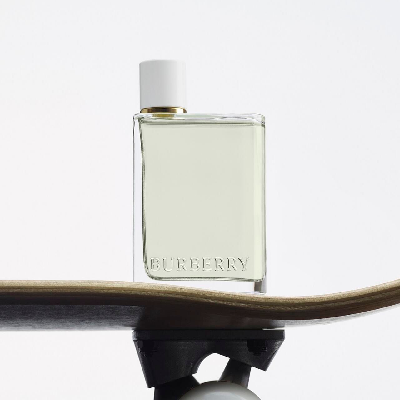 Flacone Burberry Her su uno skateboard. Forma quadrata, tappo bianco, colletto dorato. Logo Burberry.