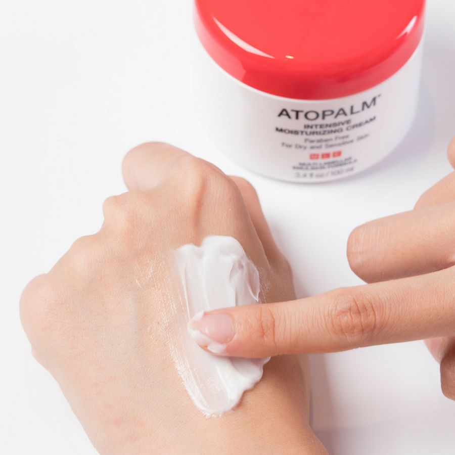 Mano con crema. Scritta: ATOPALM INTENSIVE MOISTURIZING CREAM. Crema applicata sulla mano. Vasetto bianco con coperchio rosso sullo sfondo.