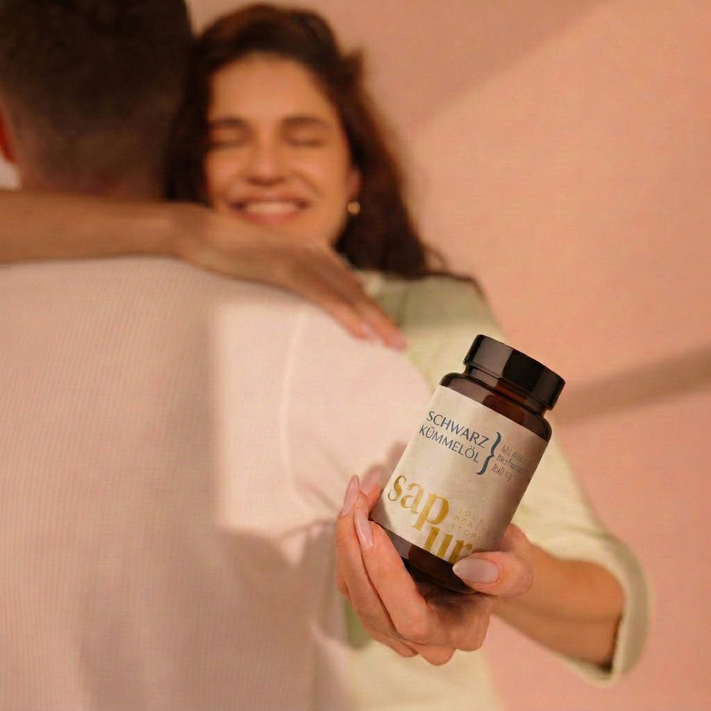 Donna abbraccia uomo, tiene flacone marrone con capsule. Scritta: SCHWARZKÜMMELÖL, Kaltgepresst & Naturbelassen, sapura.