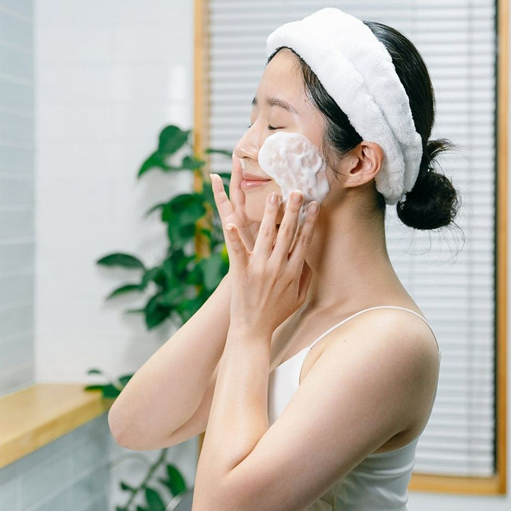 Donna con fascia bianca, applica schiuma sul viso. Mani sul viso. Sfondo: bagno.