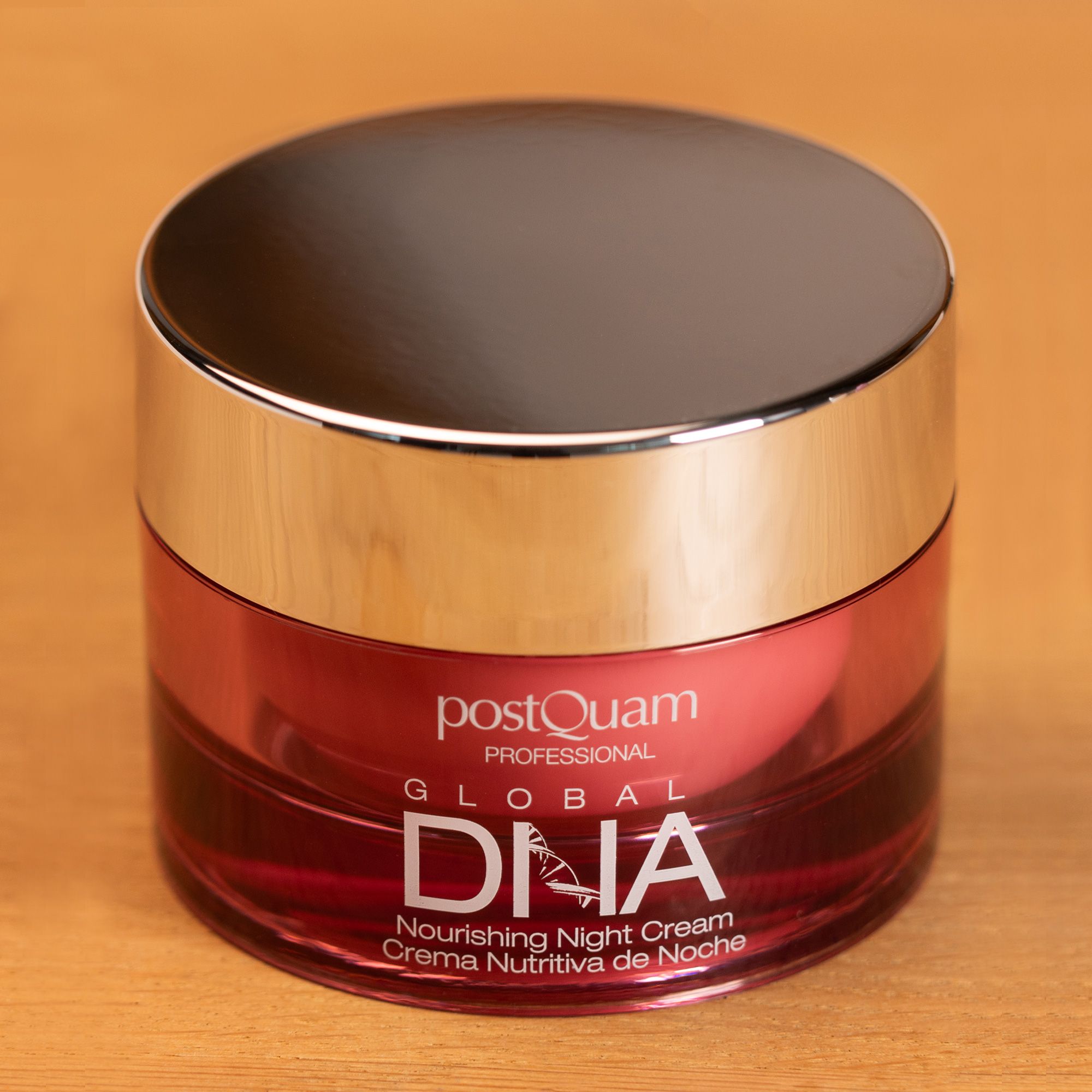 Vaso con coperchio argentato. Scritta: Postquam Global DNA Nourishing Night Cream. Crema rosa nel vetro, su fondo in legno.